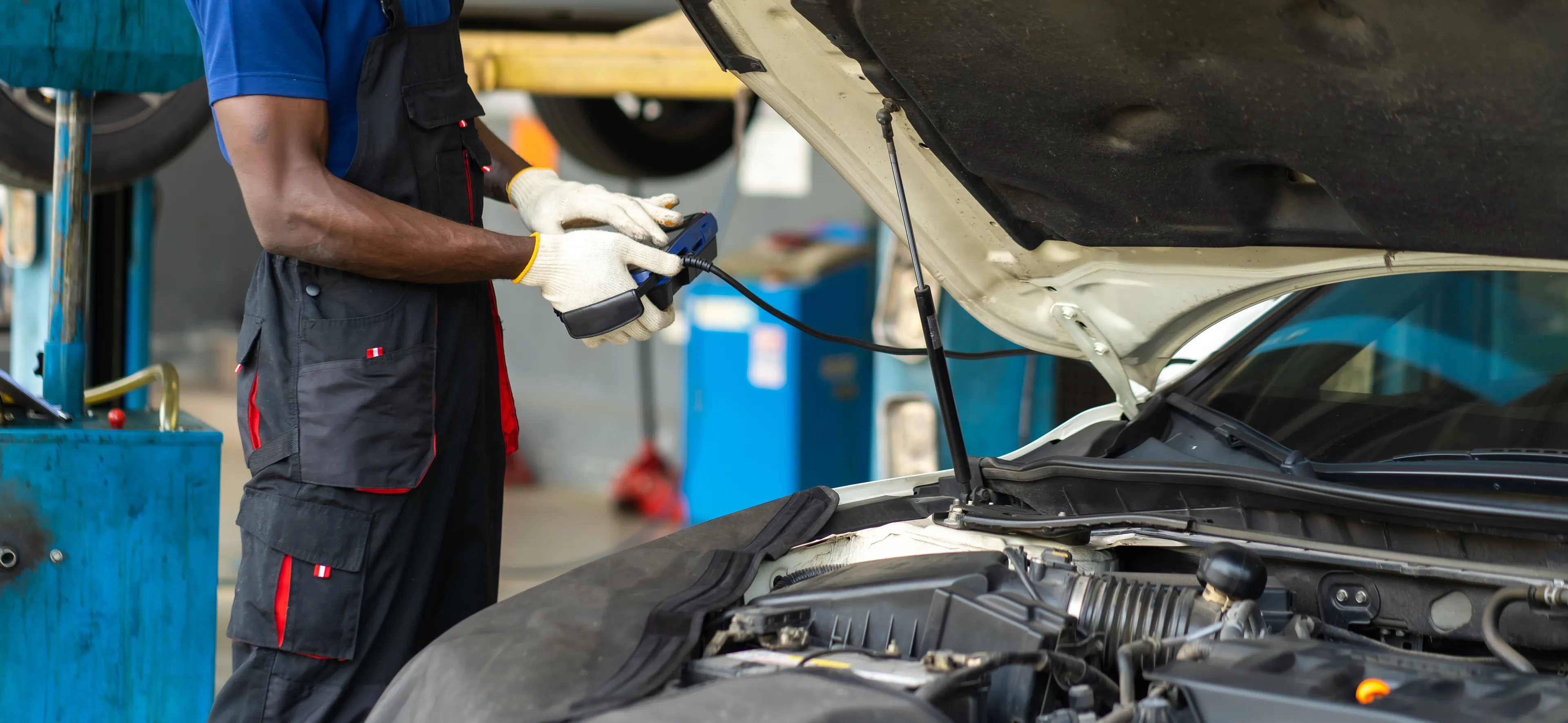 Mechanic using a diagnostic scanner on a car with the hood open in an auto repair shop.