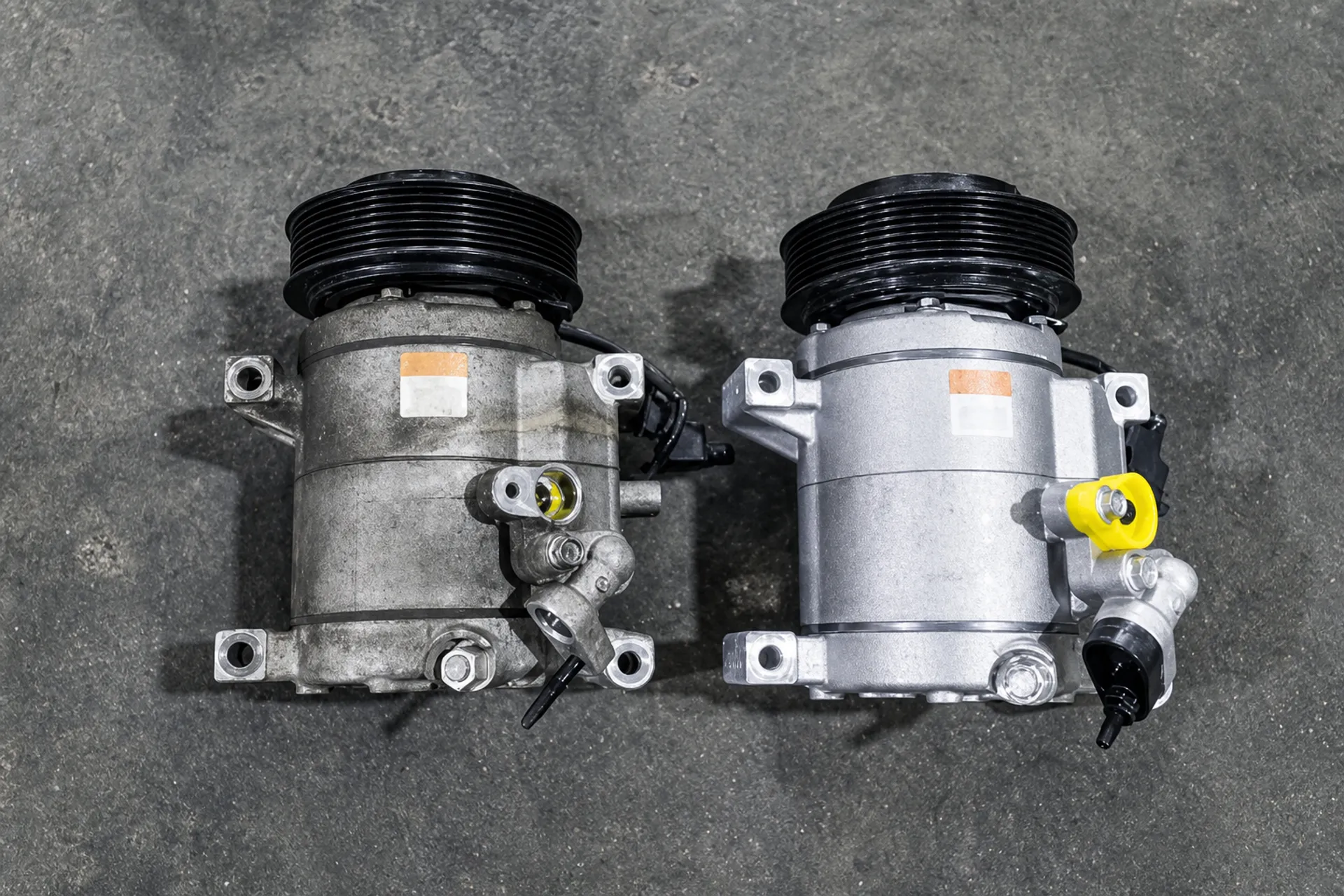 Old and new automotive AC compressors displayed side by side on a workshop floor.